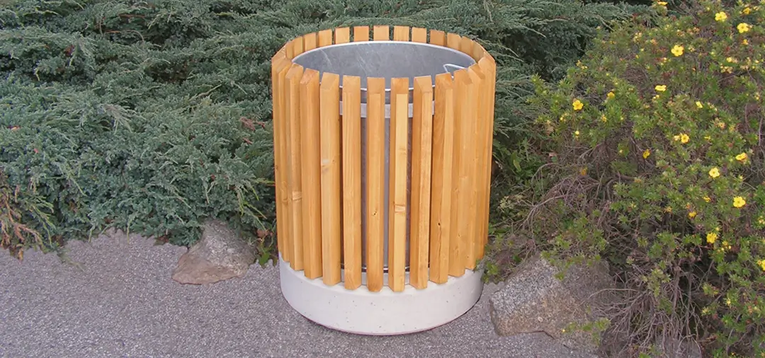 Runder Abfallbehälter LEEDS mit vertikalen Holzlatten aus Fichte und grauem Betonsockel, steht auf Kiesfläche neben Büschen.