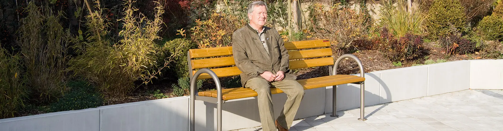 Älterer Mann sitzt auf der Seniorenbank SAN REMO mit Metallarmlehnen im Freien, umgeben von herbstlicher Bepflanzung.