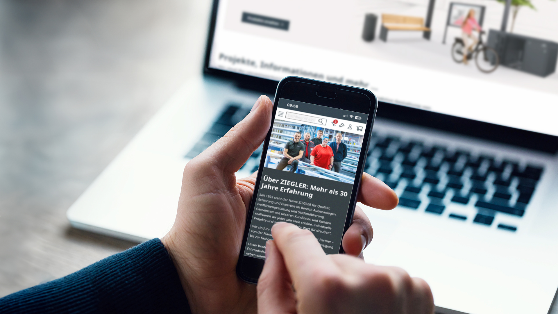 Person hält Handy in der Hand, im Hintergrund befindet sich ein Laptop