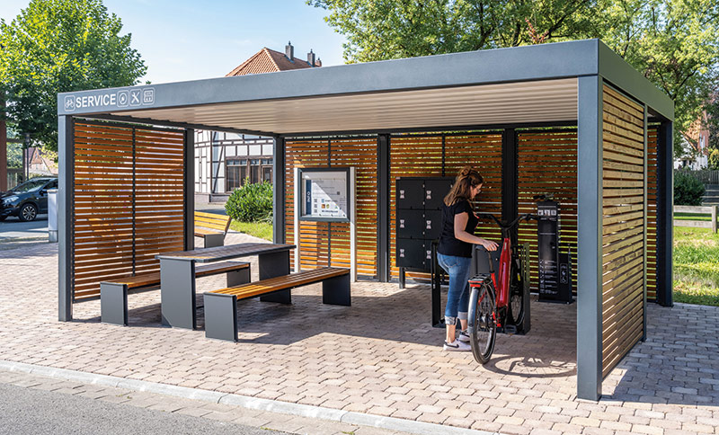 Überdachte Fahrradabstellanlage mit Sitzbank, Tisch und abschließbaren Fahrradboxen, umgeben von Holzlamellen und Metallrahmen