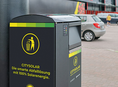 Moderner Abfallbehälter mit Aufschrift 'CITYSOLAR Die smarte Abfalllösung mit 100% Solarenergie' und Piktogramm einer Person, die Müll in einen Korb wirft.