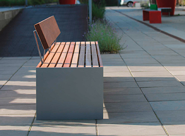 Moderne Sitzbank mit Holzlatten und grauem Betonsockel auf gepflastertem Platz mit roten Sitzwürfeln im Hintergrund.