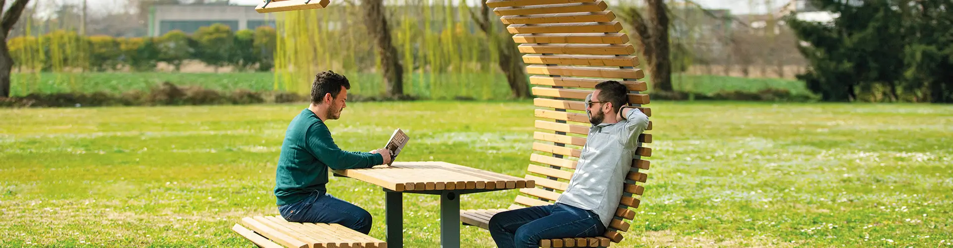 Zwei Männer sitzen an einem Tisch mit geschwungener Holzüberdachung im Freien auf einer Wiese.