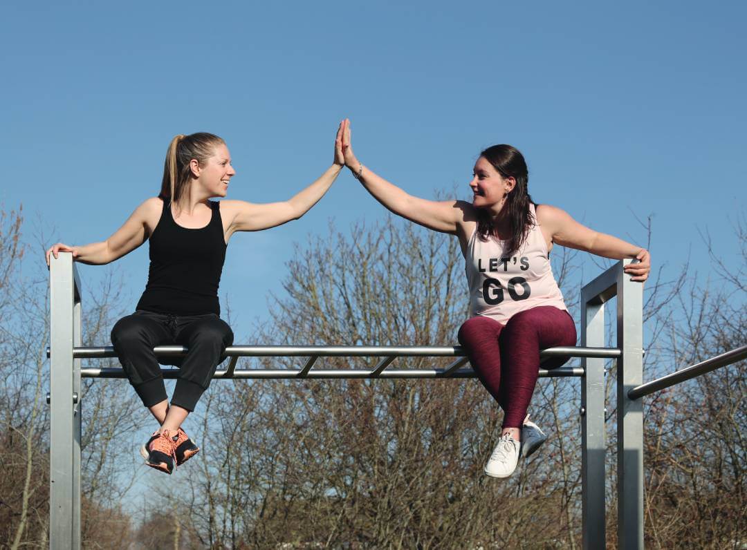 Zwei junge Frauen sitzen auf einem Sportgerät und klatschen sich ab