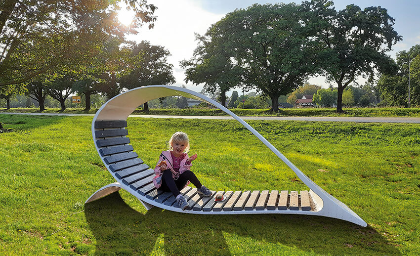 Kind sitzt auf einer geschwungenen Liegebank aus Holz und Metall auf einer Wiese mit Bäumen im Hintergrund