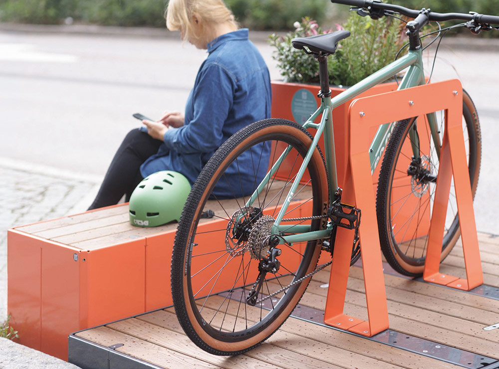 Fahrrad steht an oranger Fahrradhalterung neben Sitzinsel aus Holz, Person sitzt auf Bank und nutzt Smartphone, grüner Fahrradhelm liegt auf Bank