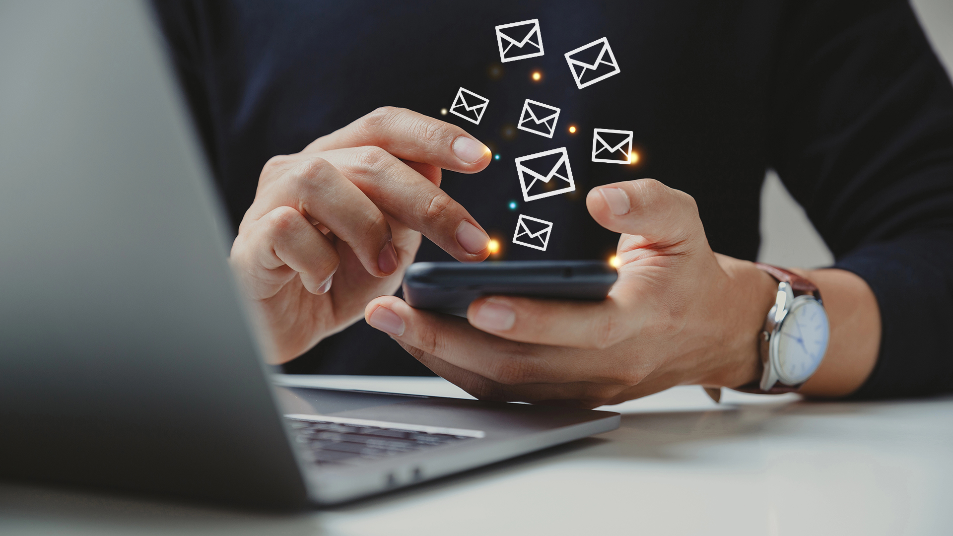 Person sitzt am Tisch und hält ein Handy in der Hand, aus welchem kleine Briefumschläge herausfliegen, vor ihr ist ein Laptop geöffnet