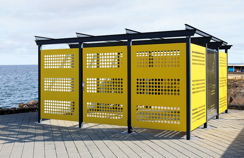Gelbe Wertstoffumhausung mit Lochmuster und schwarzem Metallrahmen auf gepflastertem Boden am Meer