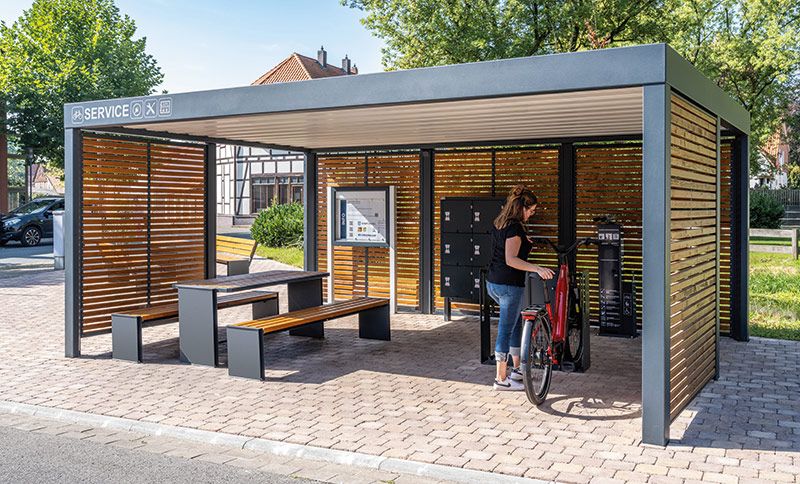Überdachte Fahrradabstellanlage mit Sitzbank, Tisch und abschließbaren Fahrradboxen, umgeben von Holzlamellen und Metallrahmen