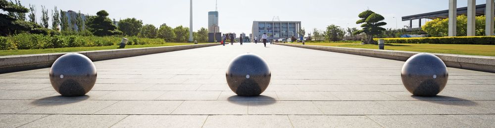 Drei kugelförmige Poller aus rekonstituiertem Marmor auf einer gepflasterten Fläche mit Gebäuden und Personen im Hintergrund