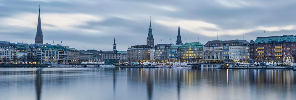 Panoramaaufnahme der Hamburger Innenstadt mit mehreren Kirchtürmen und Gebäuden am Wasser bei bewölktem Himmel.