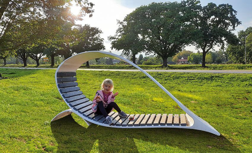 Kind sitzt auf einer geschwungenen Liegebank aus Holz und Metall auf einer Wiese mit Bäumen im Hintergrund