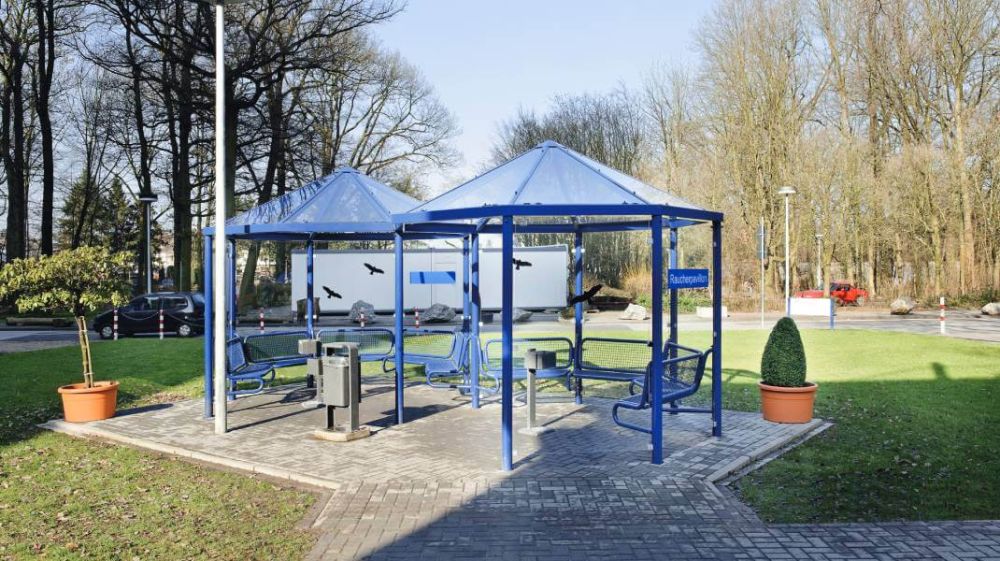 Blauer Raucherunterstand steht in der MItte eines kleinen Parkes